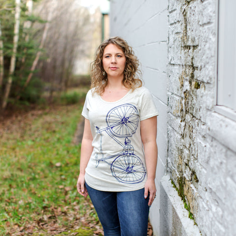 women in oatmeal heathered scoop neck t-shirt with screen printed purple bicycle standing outside next to mossy gray brick building and trees