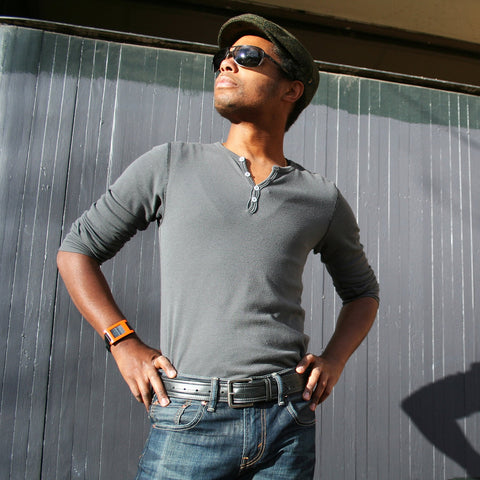 Man standing looking into sun wearing sunglasses, hat, gray shirt, jeans, and relclaim bike tube belt