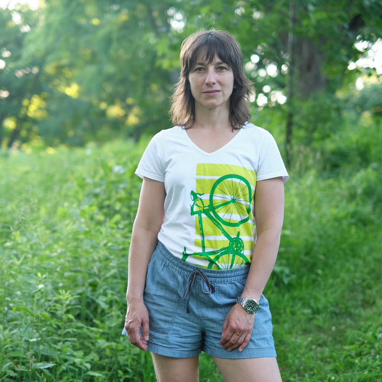female model wearing white V-neck with green printed bike shirt in front of greenery